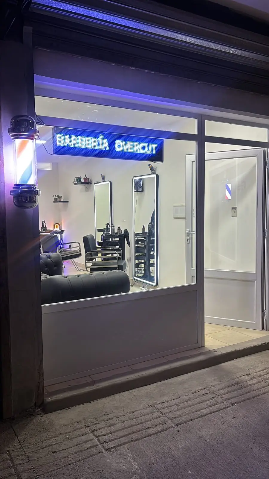 La vidriera de noche con el cartel LED «Barbería Overcut» y el poste de barbero encendido